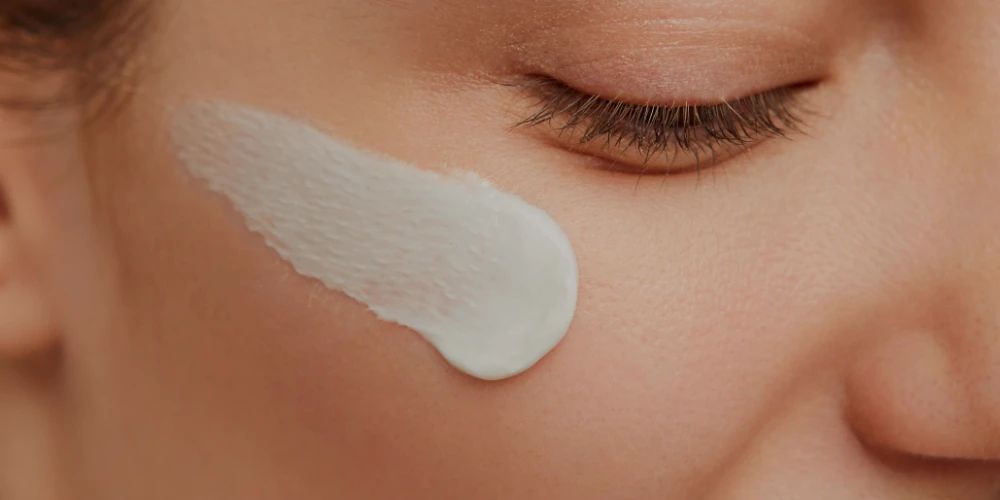 Close-up of a woman's face with a streak of white cream on her cheek, near a closed eye. The skin appears smooth, conveying a sense of skincare routine.